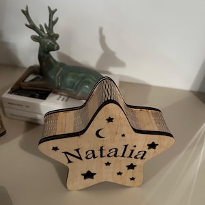 May include: A wooden star-shaped box with the name "Natalia" engraved on it. The box has a crescent moon and star cutouts. The box is made of light-colored wood and is sitting on a light-colored surface.