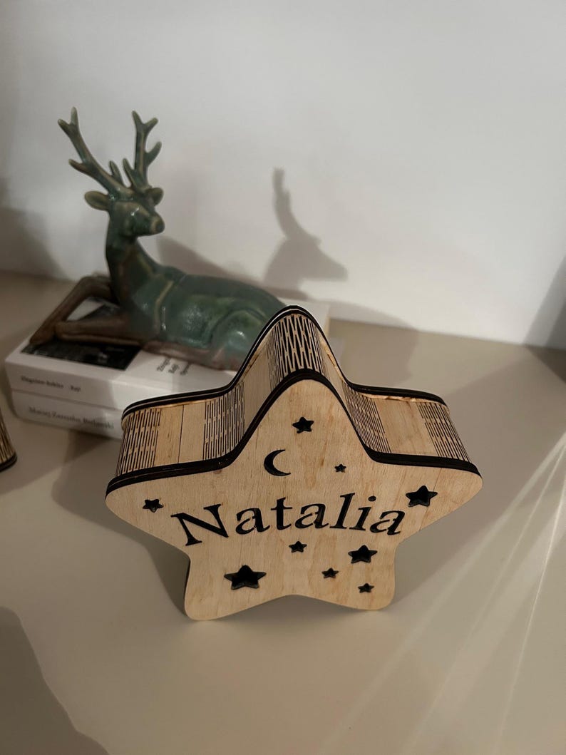 May include: A wooden star-shaped box with the name "Natalia" engraved on it. The box has a crescent moon and star cutouts. The box is made of light-colored wood and is sitting on a light-colored surface.