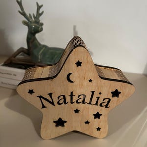 May include: Wooden star-shaped box with the name "Natalia" engraved in a bold font. The box is decorated with small star cutouts and a crescent moon. The box is made of light-colored wood.