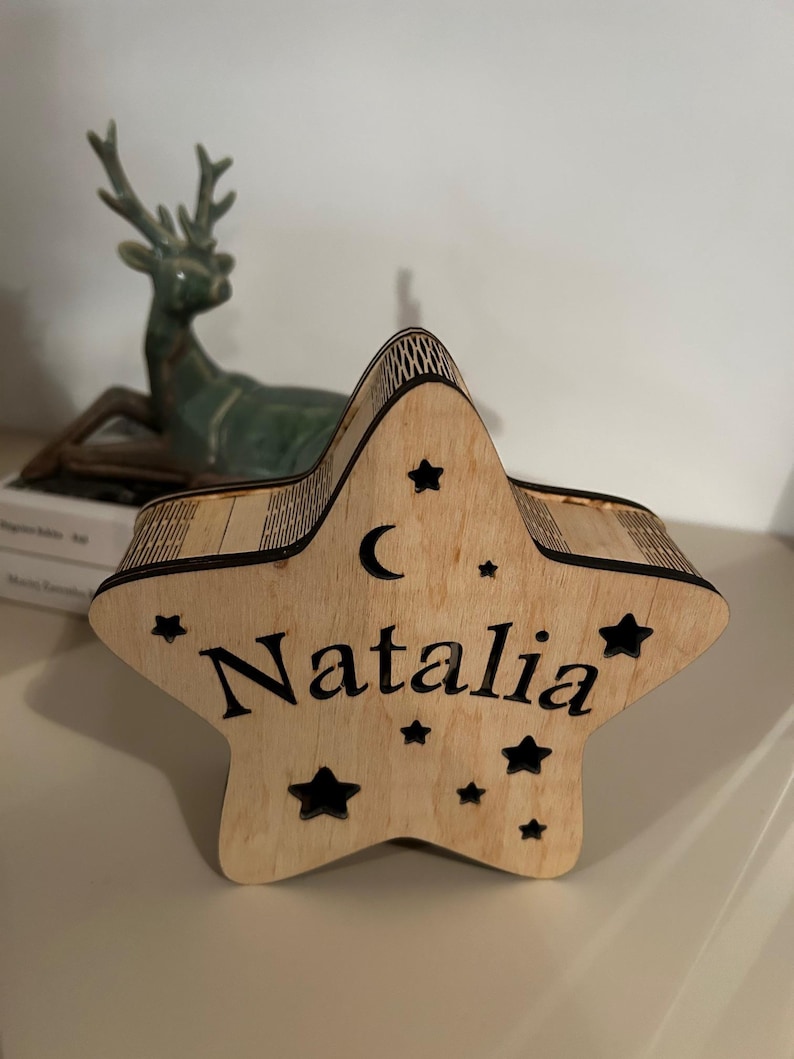 May include: Wooden star-shaped box with the name "Natalia" engraved in a bold font. The box is decorated with small star cutouts and a crescent moon. The box is made of light-colored wood.