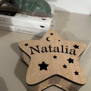 May include: A wooden star-shaped box with the name "Natalia" engraved on the top. The box features a crescent moon and star cutouts. The box is light brown with black accents.