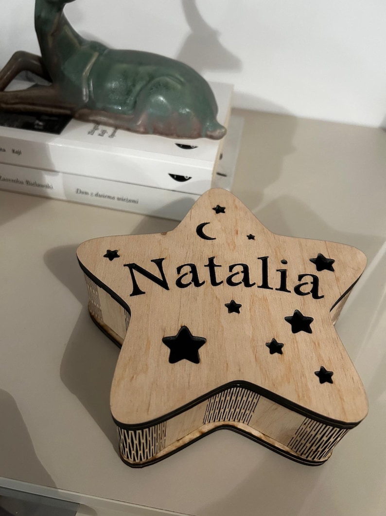 May include: A wooden star-shaped box with the name "Natalia" engraved on the top. The box features a crescent moon and star cutouts. The box is light brown with black accents.
