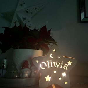 May include: A wooden star-shaped night light with the name "Oliwia" cut out and illuminated. The light features star and crescent moon cutouts. A white star decoration and a poinsettia plant are in the background.