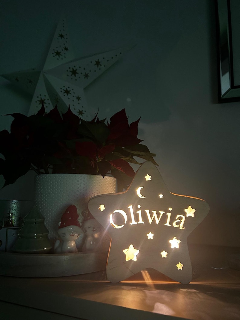 May include: A wooden star-shaped night light with the name "Oliwia" cut out and illuminated. The light features star and crescent moon cutouts. A white star decoration and a poinsettia plant are in the background.