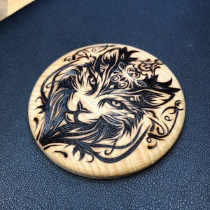 Op de afbeelding: Een ronde houten onderzetter met een gedetailleerd zwart gegraveerd ontwerp van een wolvengezicht. De onderzetter is gemaakt van lichtgekleurd hout en heeft ingewikkelde patronen rond het wolvengezicht.