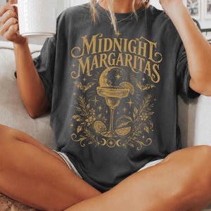 Può includere: Una t-shirt a maniche corte color grigio antracite con una stampa grafica color oro. Il design mostra la scritta "MIDNIGHT MARGARITAS" sopra un'illustrazione di un bicchiere da margarita, pipistrelli, stelle, fasi lunari e spicchi di agrumi.