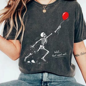 May include: A charcoal gray short-sleeve t-shirt featuring a white graphic of a running skeleton reaching for a red balloon. The text "Well... that went well." is printed in white below the skeleton. The shirt has a crew neckline and a relaxed fit.