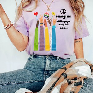 May include: A light purple t-shirt with the words "Imagine all the people living life in peace" and a peace sign. The shirt features colorful arms holding up a heart, peace sign, flower, and rock on hand gesture.