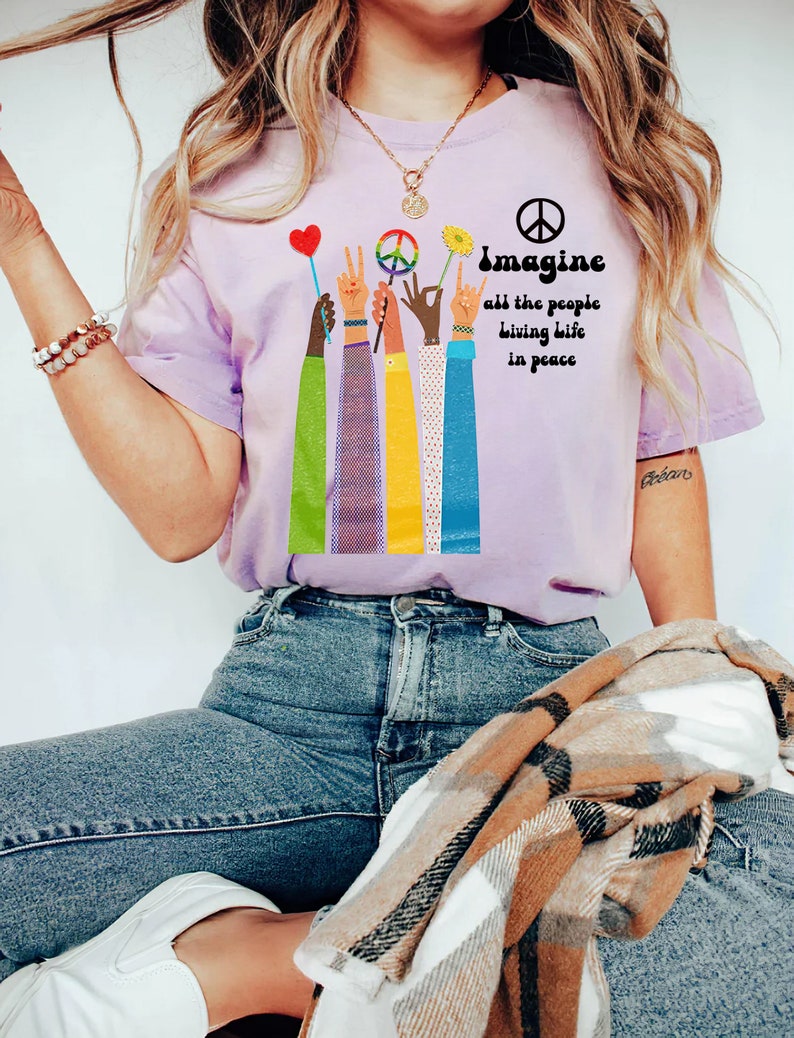 May include: A light purple t-shirt with the words "Imagine all the people living life in peace" and a peace sign. The shirt features colorful arms holding up a heart, peace sign, flower, and rock on hand gesture.