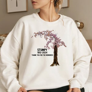 Può includere: Felpa bianca con un'immagine di un albero di ciliegio in fiore nero e rosa. Il testo "Stumpy 1912-2024 Grazie per i ricordi!" è stampato sotto l'albero.