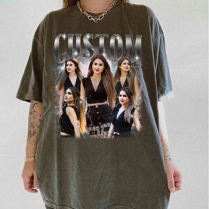 May include: Olive green t-shirt with a custom design featuring a woman in various poses, with the word "CUSTOM" in silver block letters across the top. The woman is wearing a black outfit. The design includes lightning bolt graphics.