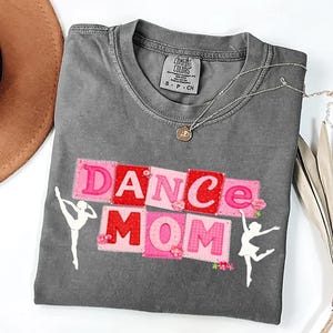 Könnte beinhalten: Graues T-Shirt mit den Worten "DANCE MOM" in rosa und roten Blockbuchstaben. Das Shirt zeigt zwei weiße Ballerina-Silhouetten und eine silberne Halskette. Das Shirt ist aus weichem Material.