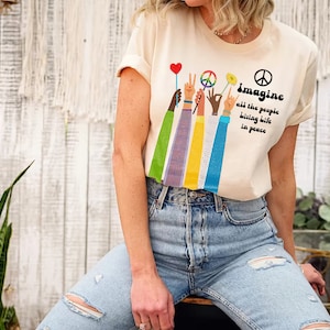 May include: Cream-colored t-shirt with the words "Imagine all the people living life in peace." The shirt features a graphic of raised hands holding a peace sign, a flower, and a heart-shaped balloon.