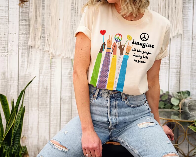 May include: Cream-colored t-shirt with the words "Imagine all the people living life in peace." The shirt features a graphic of raised hands holding a peace sign, a flower, and a heart-shaped balloon.
