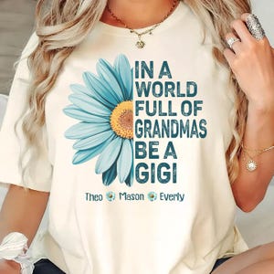 Könnte beinhalten: Weißes T-Shirt mit einem blauen Gänseblümchen-Motiv und dem Text "In einer Welt voller Omas, sei eine Gigi" mit den Namen Theo, Mason und Everly darunter.