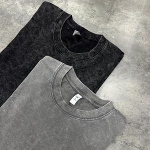 May include: Two stone-washed, crew neck t-shirts. One is dark gray, the other is a lighter gray. The shirts are folded and laid on a gray surface. The fabric has a textured, mottled appearance.