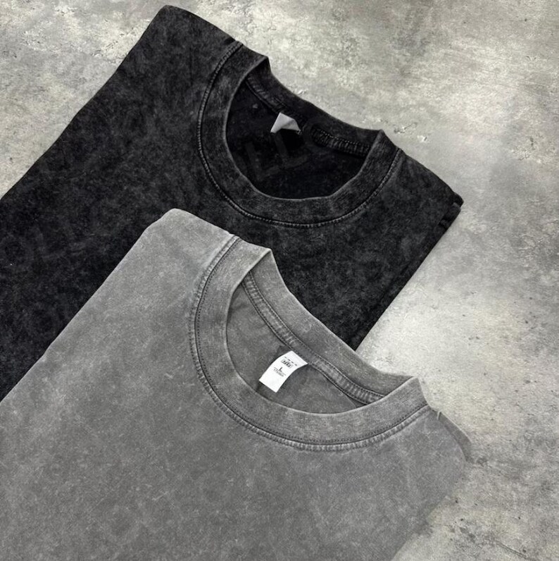 May include: Two stone-washed, crew neck t-shirts. One is dark gray, the other is a lighter gray. The shirts are folded and laid on a gray surface. The fabric has a textured, mottled appearance.