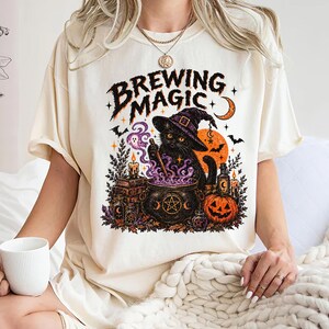 Può includere: Una t-shirt color crema con una stampa grafica di un gatto nero con un cappello da strega che mescola un calderone con fumo viola. Il testo BREWING MAGIC è scritto sopra il gatto, circondato da zucche, candele, libri e pipistrelli.