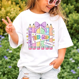 Op de afbeelding: Een wit t-shirt met een kleurrijke afbeelding van vijf potloden in paars, geel, groen, blauw en oranje. Boven een wimpel met de tekst THIRD GRADE zit een paarse ruitjesstrik en een klein label met de naam Emma eraan bevestigd.