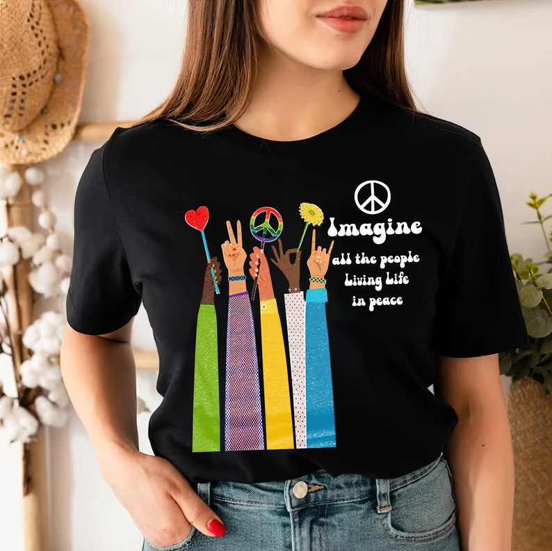 May include: Black t-shirt with a colorful graphic of raised hands holding peace symbols and flowers. The text "Imagine all the people living life in peace" is printed in white. The shirt has a crew neck and short sleeves.