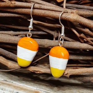 May include: A pair of dangle earrings featuring candy corn shapes. The candy corn is painted in orange, white, and yellow.