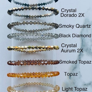 May include: A set of ten bracelets made of crystal beads in various colors, including jet black, smoky quartz, black diamond, crystal dorado, crystal aurum, smoked topaz, topaz, and light topaz. Each bracelet is stacked on top of the other, with the jet black bracelet at the top and the light topaz bracelet at the bottom. The bracelets are labeled with their corresponding colors.