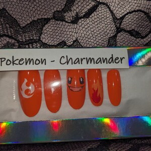 May include: A set of orange press-on nails with Pokemon Charmander designs. The nails feature a flame design, a Charmander face, and a Pokemon symbol.