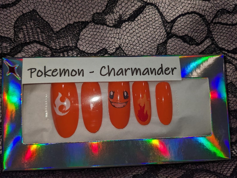 May include: A set of orange press-on nails with Pokemon Charmander designs. The nails feature a flame design, a Charmander face, and a Pokemon symbol.