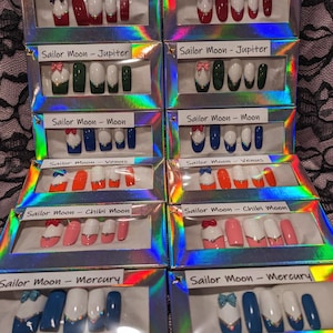 May include: Ten boxes of fake nails with different designs inspired by the Sailor Moon characters. Each box has a different color scheme and design, including red, blue, green, pink, and white. The boxes are labeled with the character's name, such as "Sailor Moon - Mars" and "Sailor Moon - Mercury".