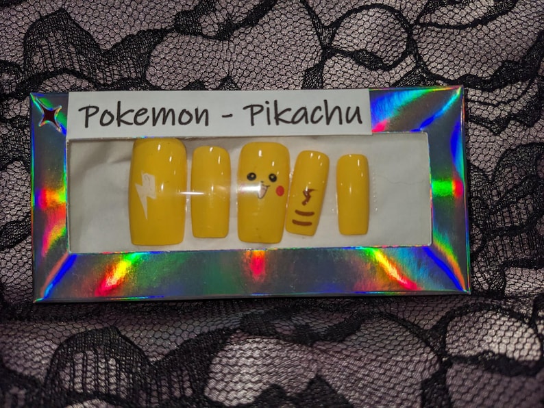 May include: A set of yellow press-on nails with a Pikachu theme. The nails feature a lightning bolt, Pikachu's face, and a tail.