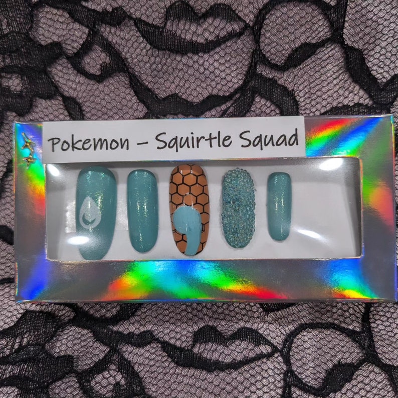 May include: A set of six false nails with a Pokemon Squirtle Squad theme. The nails are in a clear plastic box with a holographic background. The nails are decorated with blue, brown, and white designs.