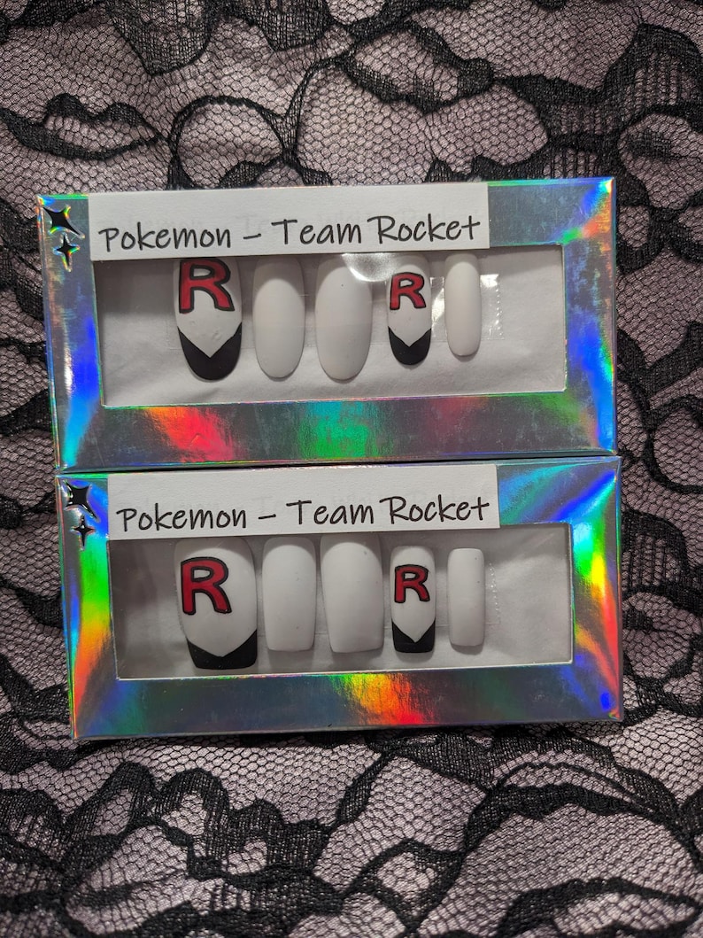 May include: Two sets of fake nails in a clear plastic box with a holographic finish. Each set has five nails with a white base and a black and red "R" design. The box has the text "Pokemon - Team Rocket" printed on it.