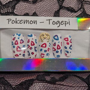 May include: A set of five false nails with a Pokemon Togepi design. The nails are white with red and blue geometric shapes and a Togepi character on the middle nail.