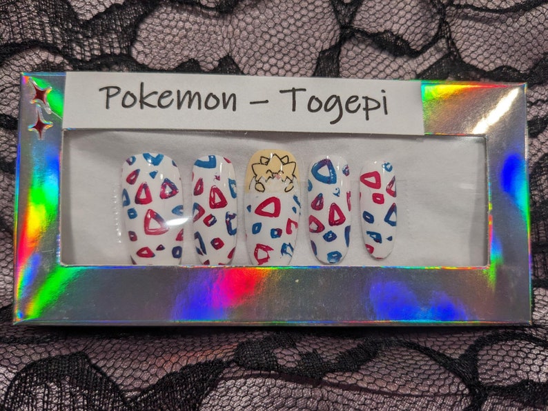 May include: A set of five false nails with a Pokemon Togepi design. The nails are white with red and blue geometric shapes and a Togepi character on the middle nail.