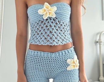 May include: A light blue crochet two-piece set. The top is a strapless bandeau style with a floral accent. The shorts are high-waisted with a matching floral accent. The set is handmade and perfect for summer.