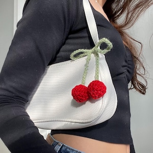 May include: White faux leather handbag with a crocheted cherry charm attached to the strap. The cherry is red with green leaves.