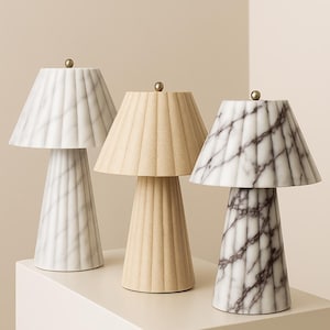 May include: Three decorative table lamps with conical bases and pleated shades. The lamps feature marble and beige finishes, topped with small gold-colored finials. The lamps are displayed on a white surface against a neutral background.