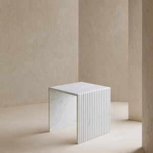May include: A white marble side table with a square top and a fluted, vertical-lined design on one side. The table is set against a neutral-toned background, showcasing its minimalist aesthetic. The table's dimensions are approximately 18 inches in height and width.