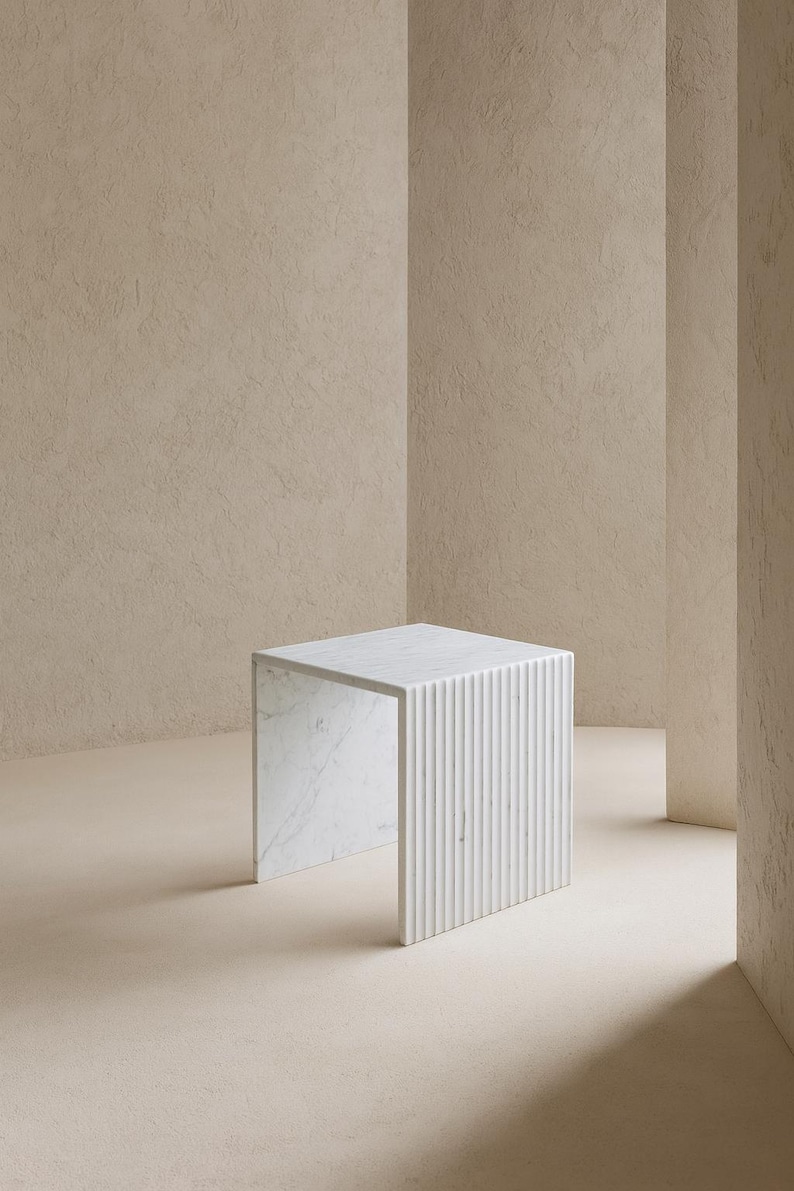 May include: A white marble side table with a square top and a fluted, vertical-lined design on one side. The table is set against a neutral-toned background, showcasing its minimalist aesthetic. The table's dimensions are approximately 18 inches in height and width.
