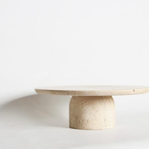 May include: A beige, round, stone cake stand with a cylindrical base. The top is a flat, circular surface, and the base is a solid, rounded column. The stand is made of a light-colored stone material, and it is set against a white background.