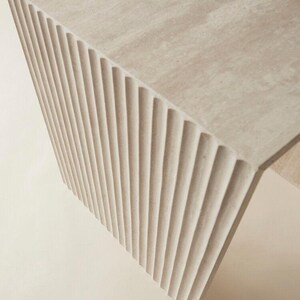 May include: A light beige, modern-style table with a textured surface and a fluted, ridged side. The table's design features a unique architectural element, showcasing a blend of form and function. The neutral color palette makes it versatile for various interior settings.