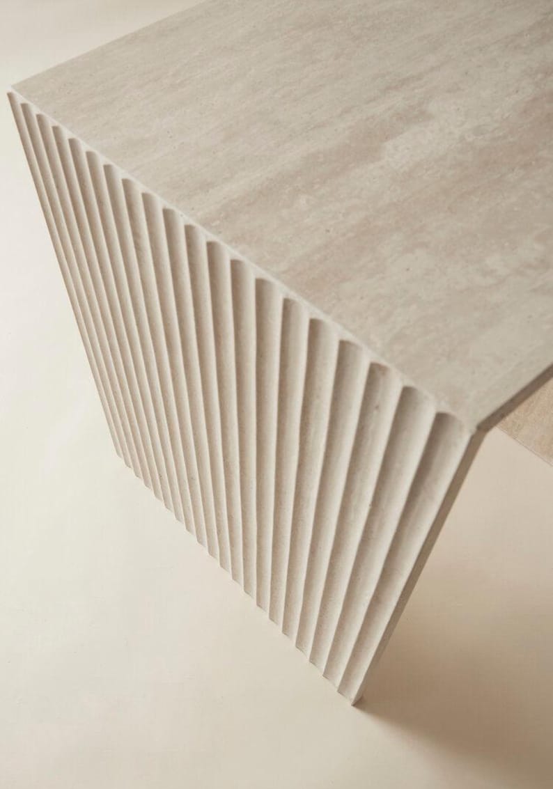 May include: A light beige, modern-style table with a textured surface and a fluted, ridged side. The table's design features a unique architectural element, showcasing a blend of form and function. The neutral color palette makes it versatile for various interior settings.
