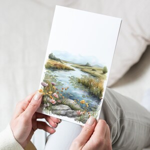 以下が含まれることがあります： 緑の草、野花、スイレンが咲く草原を流れる小川を描いた水彩画。空は白雲のある薄い青色です。