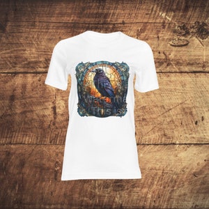 K&ouml;nnte beinhalten: Wei&szlig;es T-Shirt mit einem grafischen Druck eines schwarzen Rabens, der auf einem Ast in einem Buntglasfensterrahmen sitzt. Das Fenster hat einen Sonnenuntergangs-Hintergrund mit blauen, gr&uuml;nen und goldenen Akzenten.