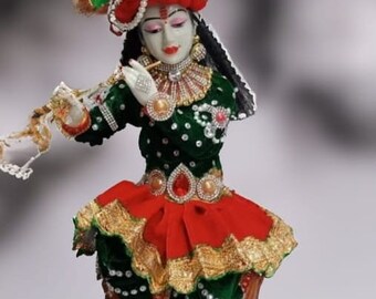 May include: A detailed figurine of a deity, adorned in a red and green outfit with gold trim. The figure wears a jeweled headdress and plays a flute. The statue is seated on a wooden base, showcasing intricate craftsmanship.