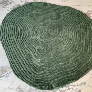 May include: An oval rug in a green colour with a concentric circle pattern. The rug is made from a soft, plush material and has a subtle texture.