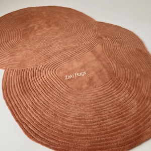 May include: A terracotta-colored rug with an irregular, organic shape featuring concentric, raised circular patterns throughout its surface. The text Zaki Rugs is printed in white in the center of the rug, which has a soft, textured pile.