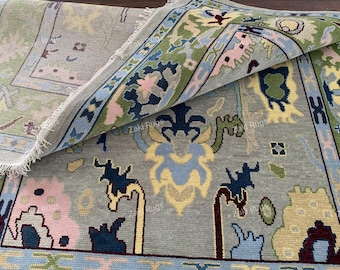 Può includere: Un tappeto arrotolato con un motivo dettagliato. Il tappeto presenta uno sfondo grigio con un bordo blu, verde e rosa. Il design include forme floreali e geometriche in vari colori.