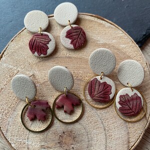May include: A set of six pairs of earrings featuring a white ceramic disc with a burgundy leaf design. Some earrings have a gold hoop with a burgundy leaf charm.