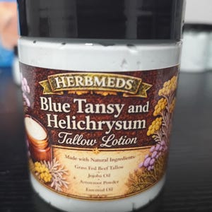 May include: A cylindrical plastic jar with a black lid containing Blue Tansy and Helichrysum Tallow Lotion by Herbmeds. The label features botanical illustrations and lists ingredients including grass fed beef tallow, jojoba oil, and arrowroot powder.